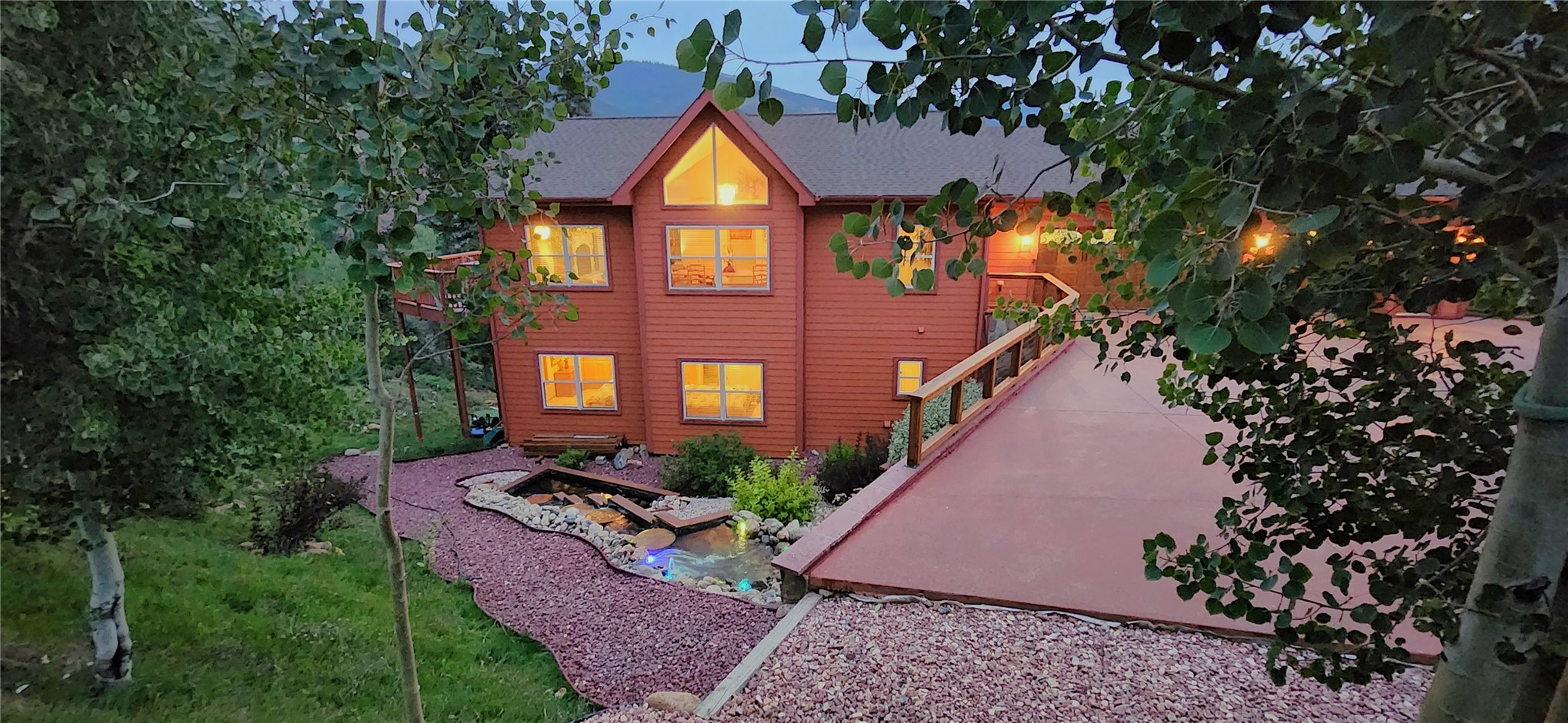 The cedar lodge glowing at twilight with warm-lit windows above a landscaped pond and waterfall, string lights strung along the deck, framed by aspen trees