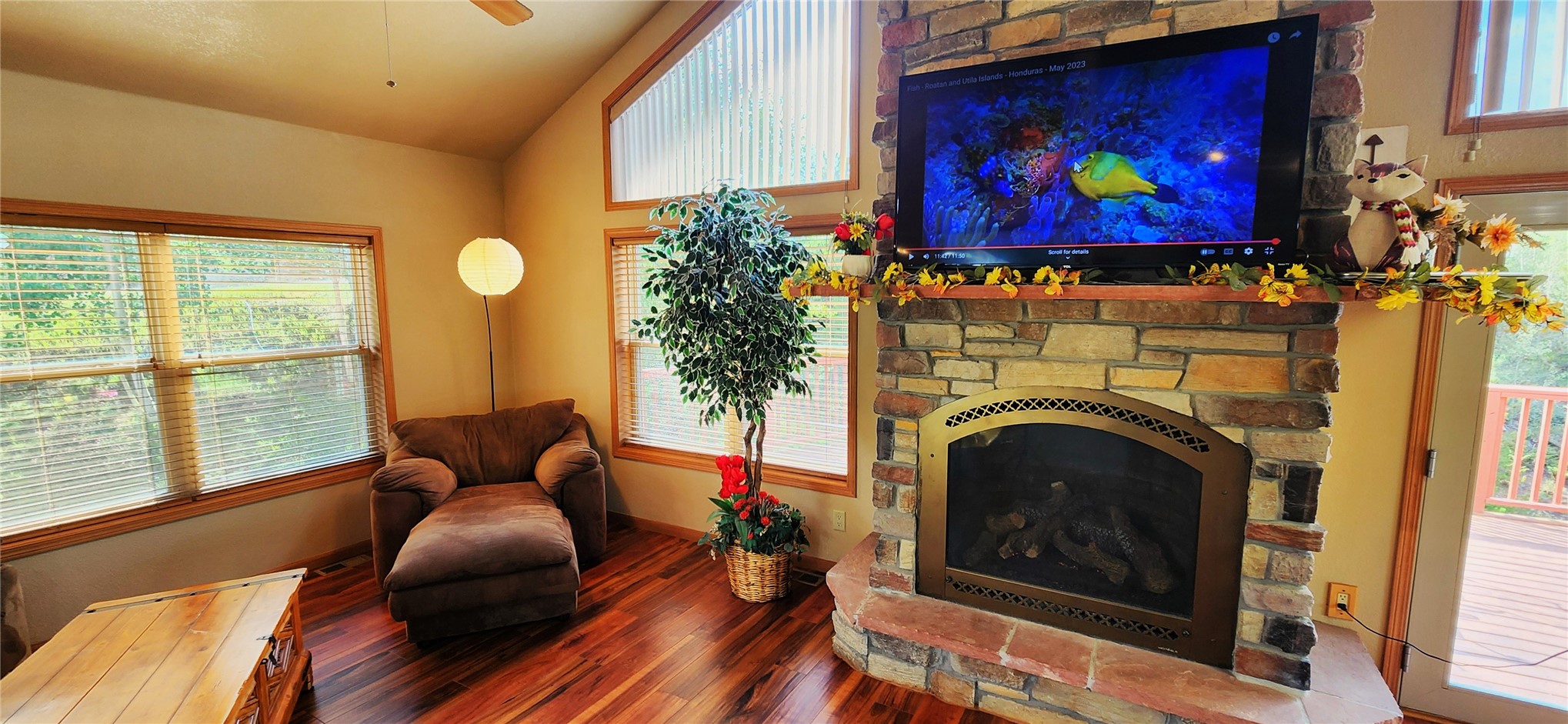 A stacked-stone gas fireplace with a wall-mounted TV above the mantel, a brown upholstered chaise in the sitting corner, and hickory floors beside the windows