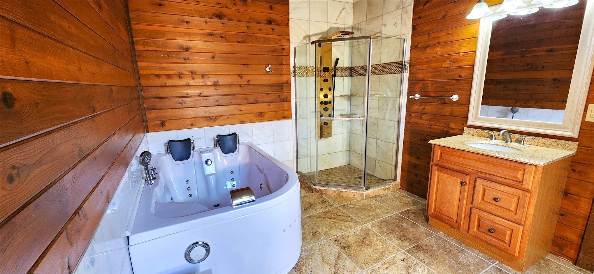The spa bathroom with a corner two-person jetted tub, a glass shower with a gold panel, cedar plank walls, and a granite-topped oak vanity