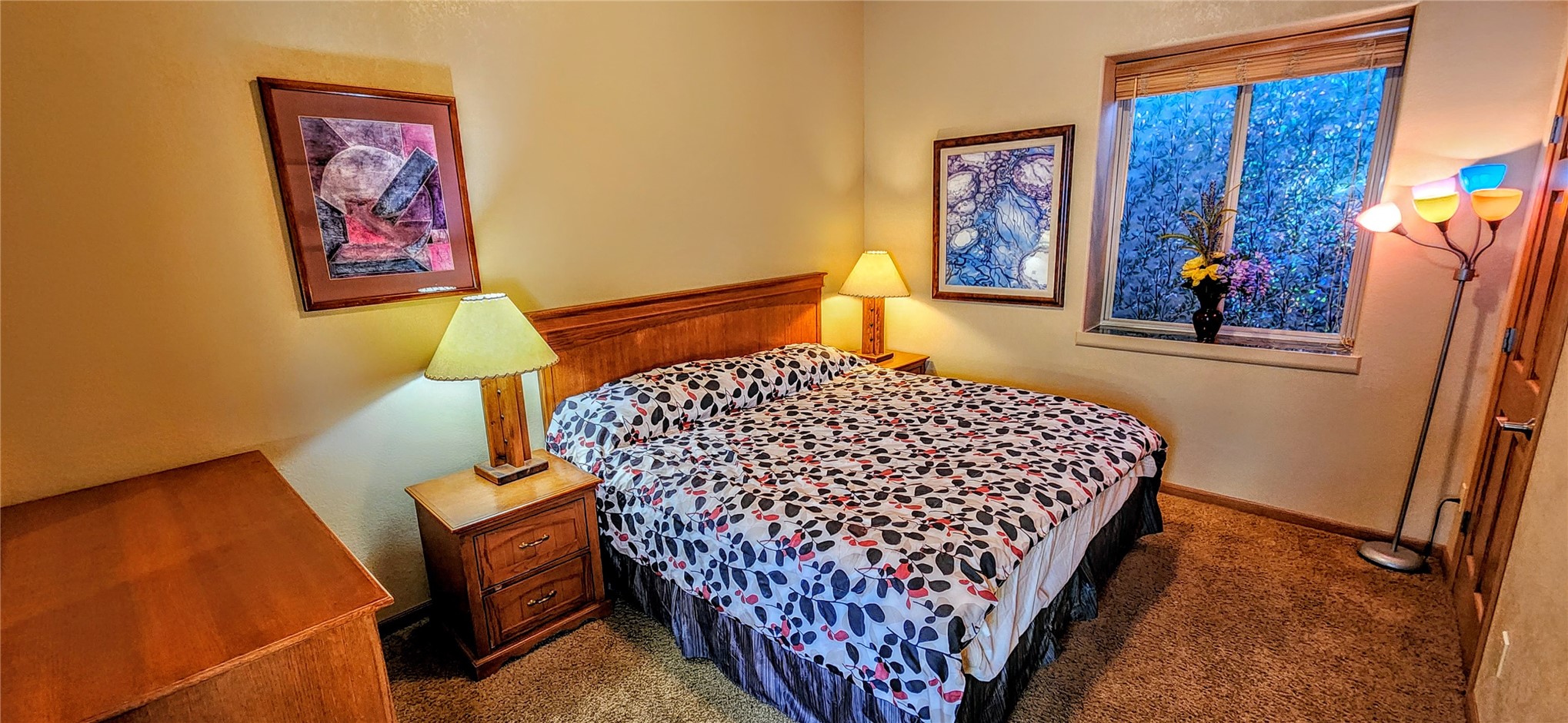 A carpeted guest bedroom with a queen bed, an oak nightstand and dresser, framed art, and a window looking into the aspens