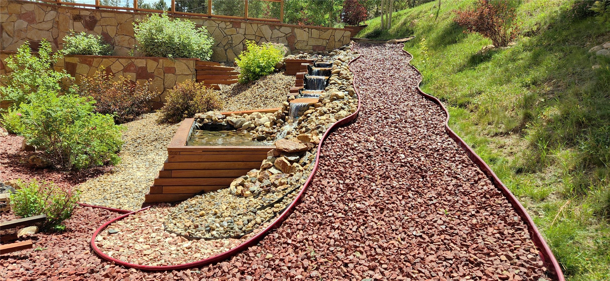 The landscaped grounds: a man-made waterfall and pond set into a stacked-stone retaining wall, with red rock paths cut along the hillside
