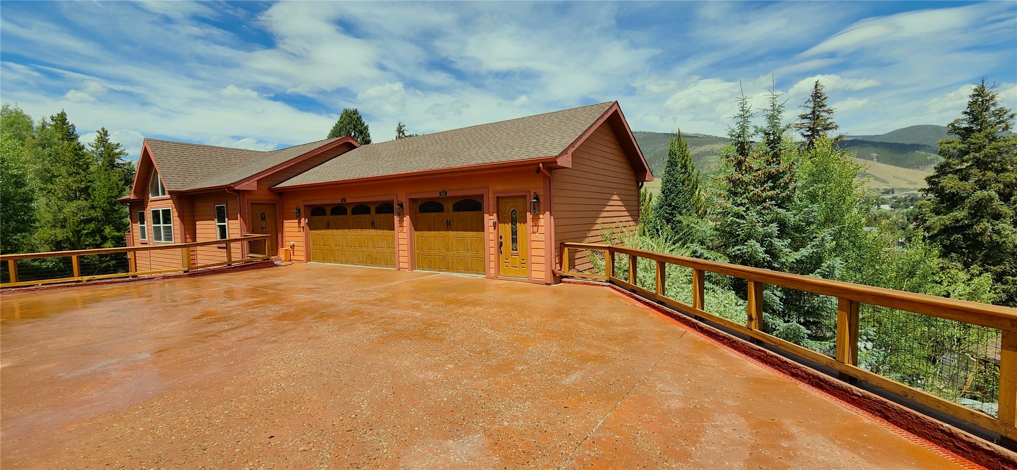 Daytime front elevation of the lodge under a blue sky, the broad terracotta driveway and deck rail in the foreground, mountains and evergreens beyond