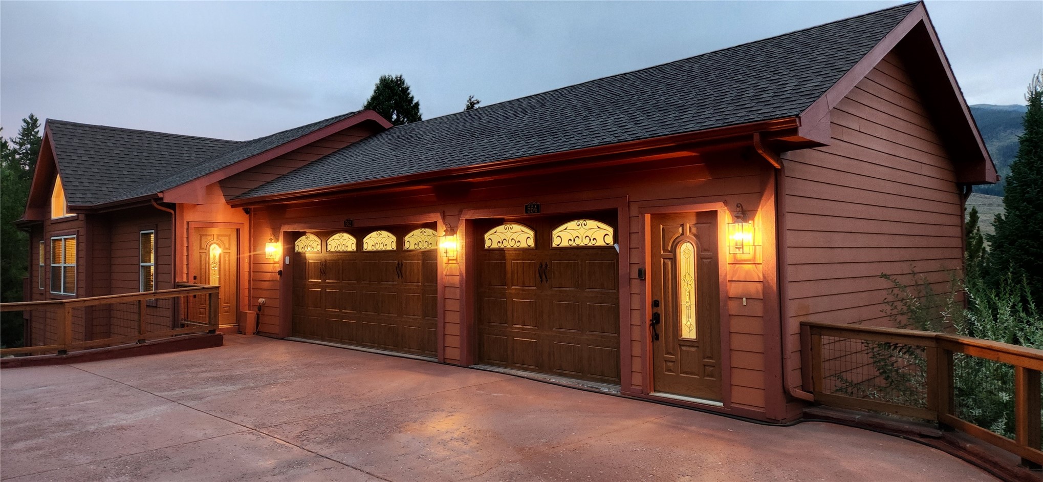 Golden-hour front elevation of 562 Piney Acres Circle, a cedar-sided mountain lodge with three carriage-style garage doors lit by sconces, a terracotta driveway, and the Tenmile Range beyond